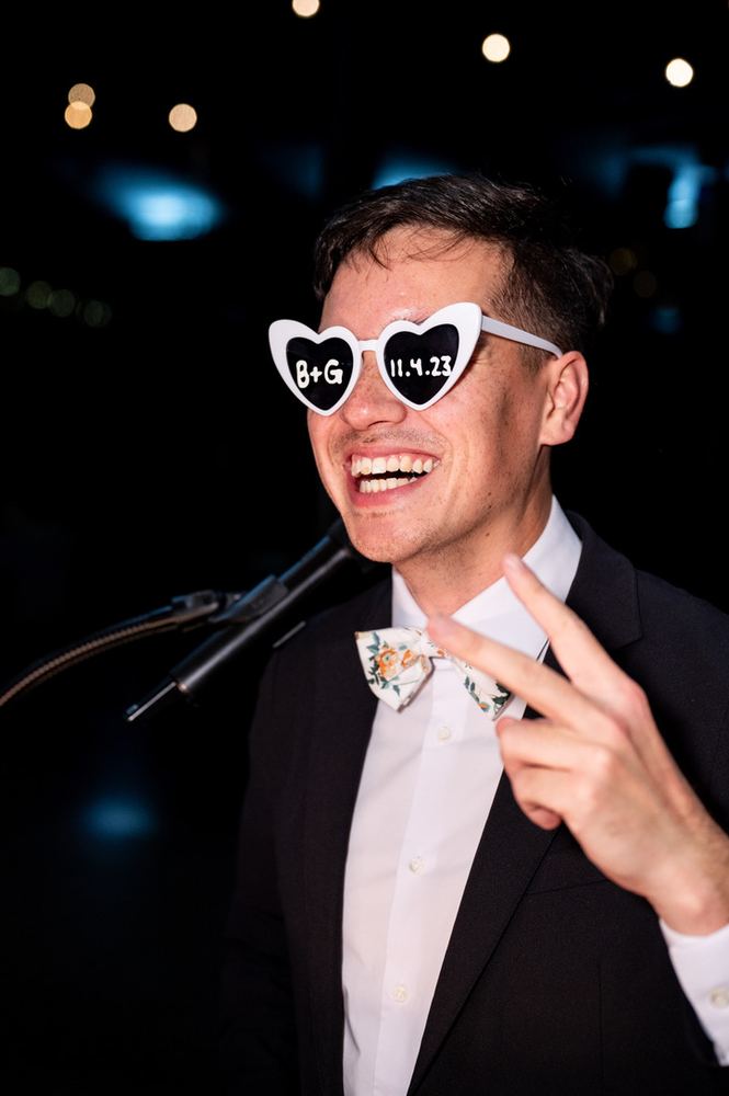 A groom in heart-shaped sunglasses laughing on the mic.